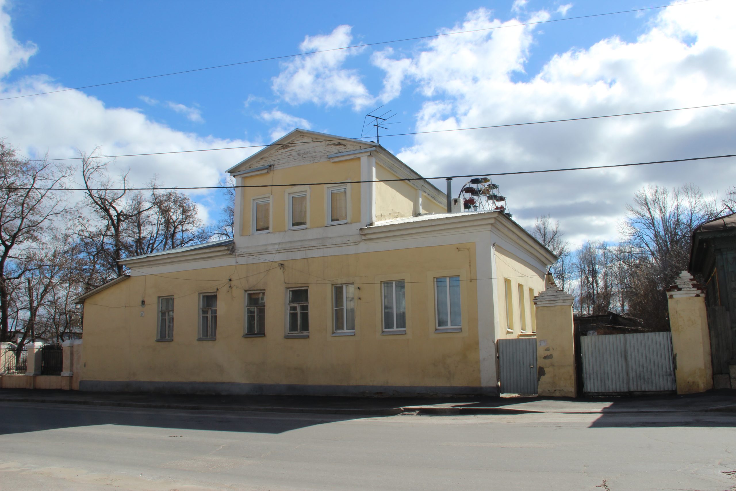 Одноэтажный жёлтый дом с трёхоконным мезонином и кирпичными воротными столбами справа на улице 9 Декабря, 21.