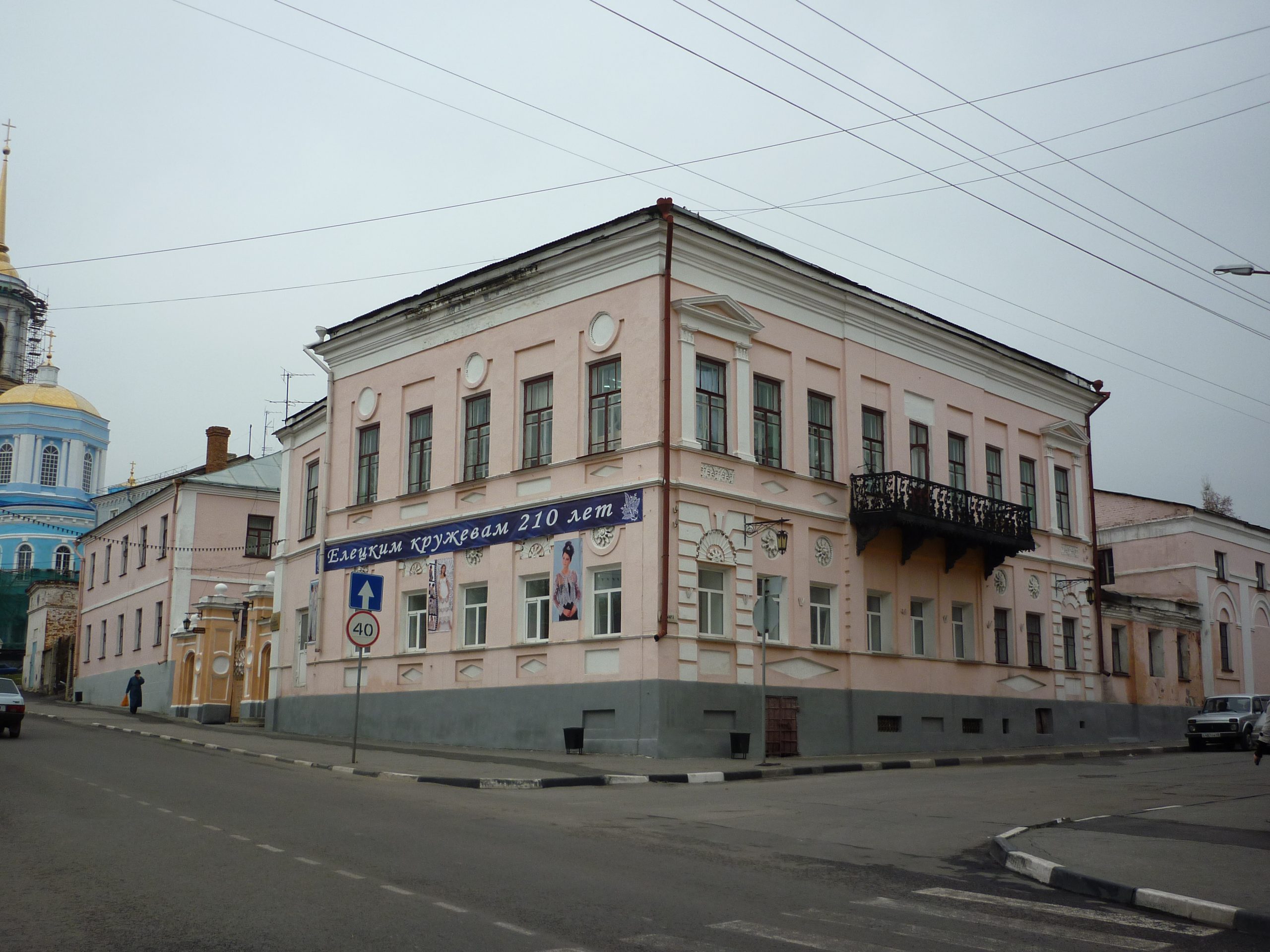 Угловой двухэтажный дом с чугунным балконом и вывеской «Елецким кружевам 210 лет»; слева видно примыкающее крыло.